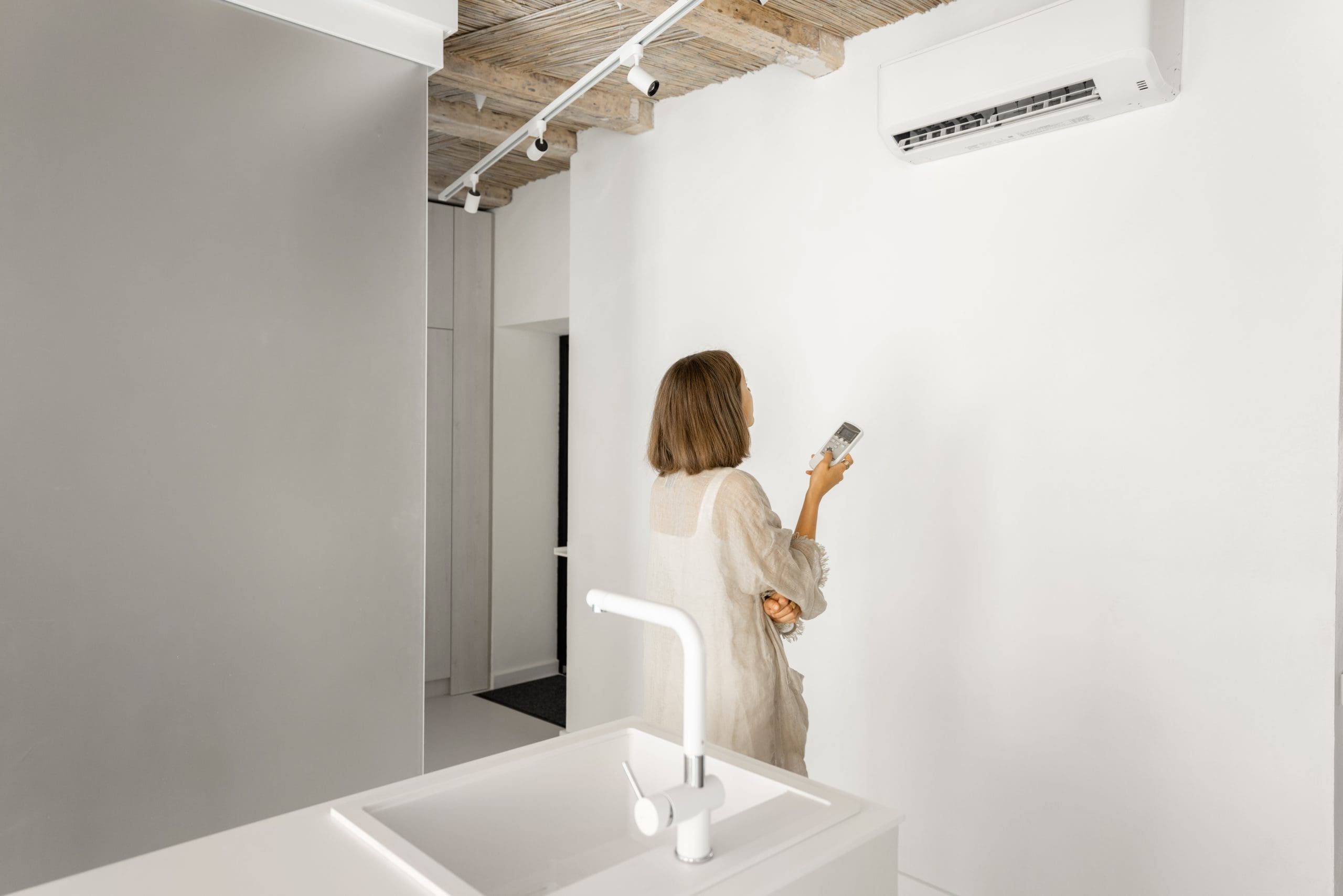 Woman sets the temperature on the air conditioner with remote control, while standing at modern white kitchen of the studio apartment